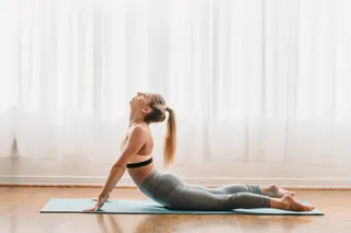 O femeie într-un suport sportiv gri execută o poziție de cobra pe un covoraș de yoga într-o cameră luminoasă cu perdele albe.