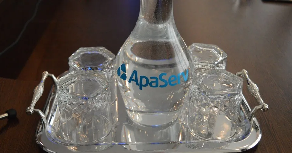 O carafă de sticlă cu apă și trei pahare de cristal sunt așezate pe o tavă metalică cu mânere decorate.