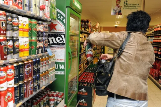 O persoană cu o geantă neagră în spate se întinde spre rafturile unui magazin din apropierea unor produse ambalate în cutii și conserve.