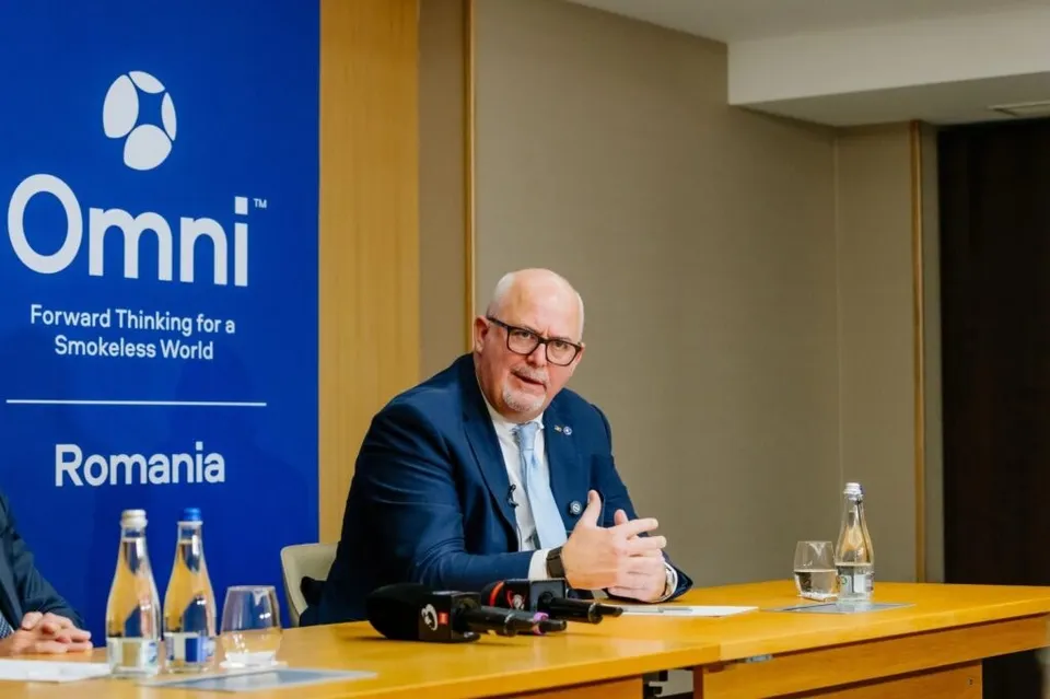 Un bărbat în costum bleumarin și cravată deschisă, cu ochelari, vorbește la o masă de lemn, cu un banner alăturat care afișează logo-ul Omni și textul 'Romania'.