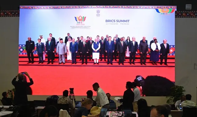 Un grup de lideri în costume oficiale stă în rând pe un podium roșu în fața unui ecran mare cu textul BRICS SUMMIT.