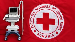 Un aparat medical alb cu ecran și tastatură este poziționat lângă un emblema circulară cu o cruce roșie pe un fundal textil roșu.