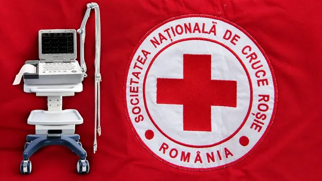 Un aparat medical alb cu ecran și tastatură este poziționat lângă un emblema circulară cu o cruce roșie pe un fundal textil roșu.
