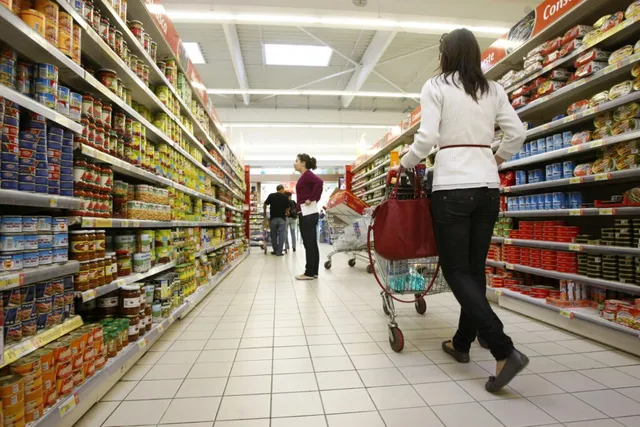 O femeie cu o geantă roșie și un cărucior de cumpărături merge pe un culoar de supermarket între rafturi pline cu produse alimentare.