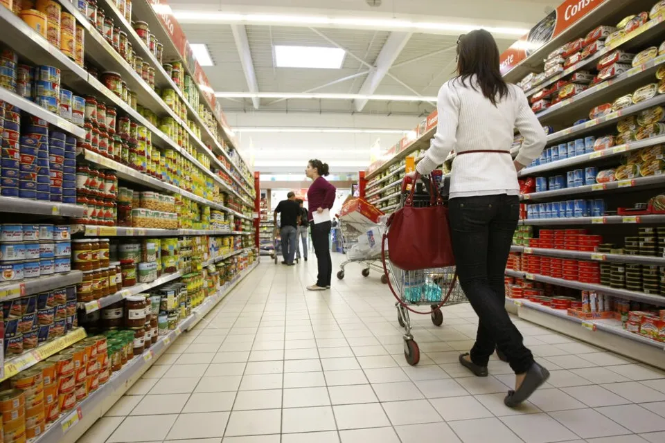 O femeie cu o geantă roșie și un cărucior de cumpărături merge pe un culoar de supermarket între rafturi pline cu produse alimentare.