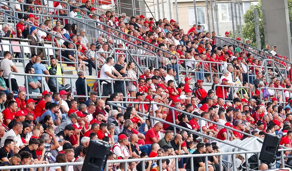 O mulțime de oameni îmbrăcați în haine roșii și albe stau pe tribunele unui stadion sub lumina soarelui.
