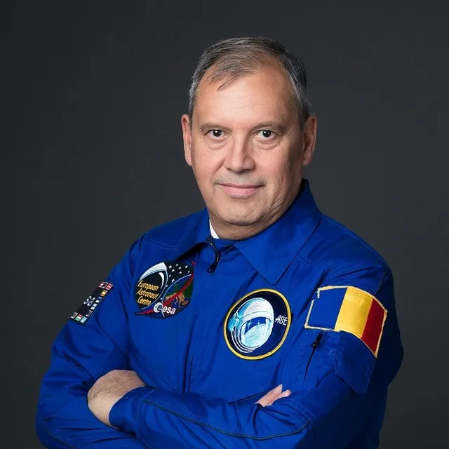 Un bărbat cu părul scurt și grij, purtând o uniformă albastră cu diverse embleme colorate, stă cu brațele încrucișate în fața unui fundal gri.