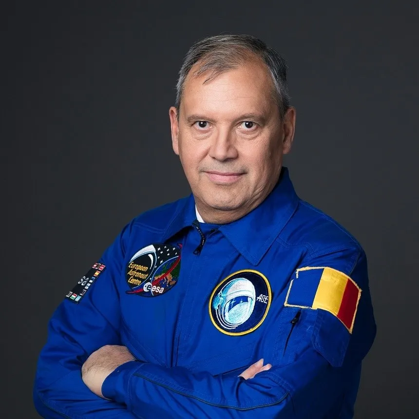 Un bărbat cu părul scurt și grij, purtând o uniformă albastră cu diverse embleme colorate, stă cu brațele încrucișate în fața unui fundal gri.