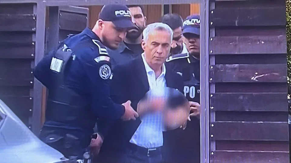 Un bărbat cu părul cărunt și cămașă albă este înconjurat de mai mulți polițiști în uniformă în fața unei porți de lemn.