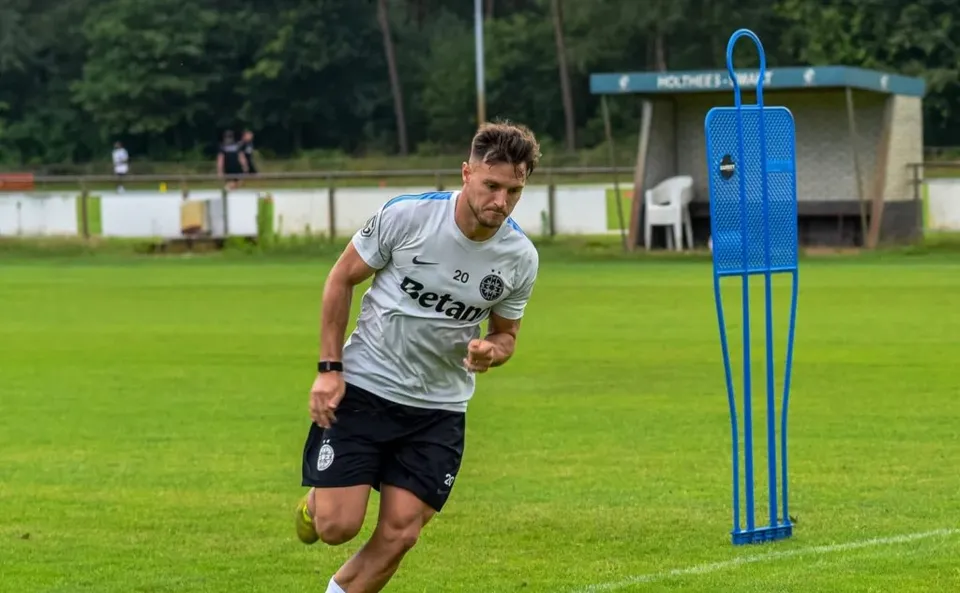 Un bărbat cu tricou alb și pantaloni scurți negri aleargă pe un teren de fotbal verde lângă un obstacol albastru.