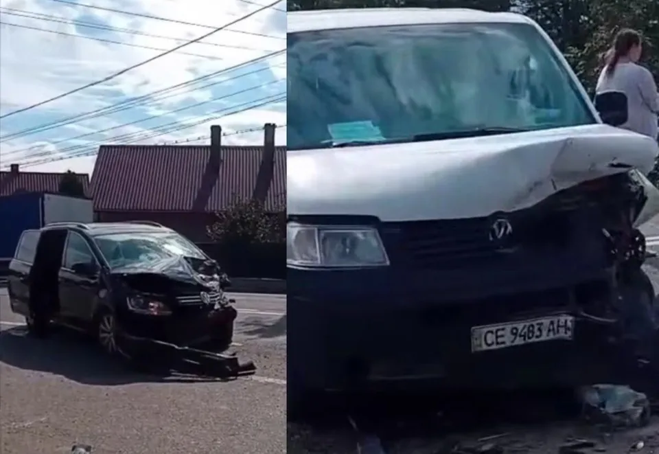 O autoutilitară albă cu partea frontală deformată este prezentată lângă o mașină neagră cu partea din față avariată pe un drum asfaltat.