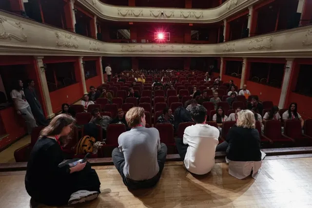 Oamenii stau pe podeaua de lemn din față, privind spre rândurile de scaune roșii ocupate într-un auditorium cu balcone decorate.