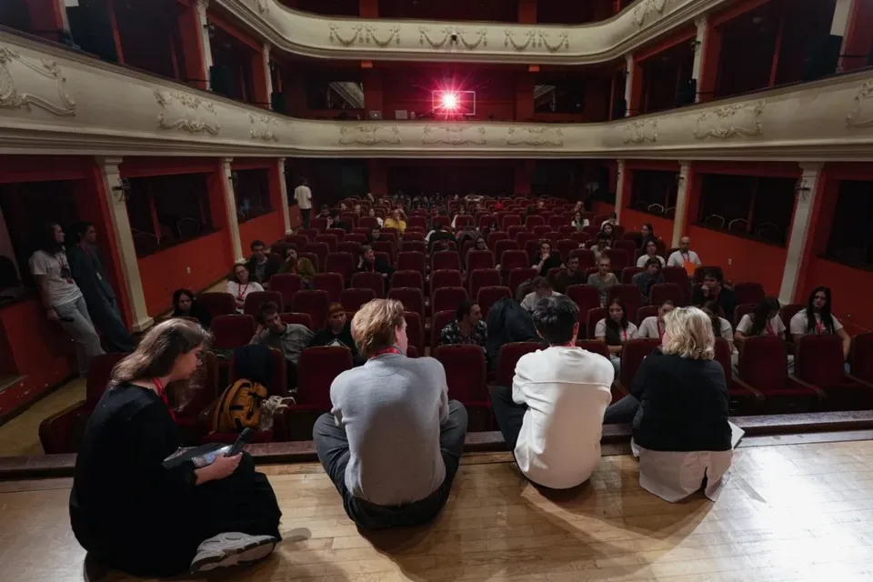 Oamenii stau pe podeaua de lemn din față, privind spre rândurile de scaune roșii ocupate într-un auditorium cu balcone decorate.