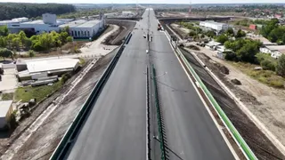 O vedere aeriană de sus a unei porțiuni de autostradă cu asfalt negru, flancată de margini verzi și zone de teren necultivat cu clădiri.
