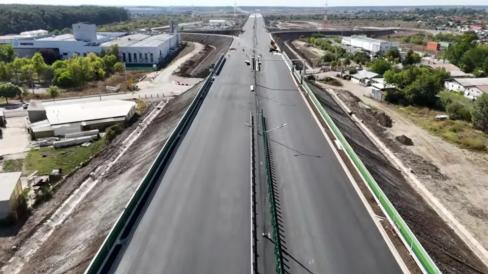 O vedere aeriană de sus a unei porțiuni de autostradă cu asfalt negru, flancată de margini verzi și zone de teren necultivat cu clădiri.