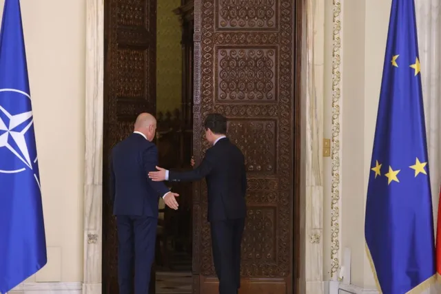 Doi bărbați în costume închise stau în fața unei uși mari de lemn, flancate de steagurile NATO și al Uniunii Europene.