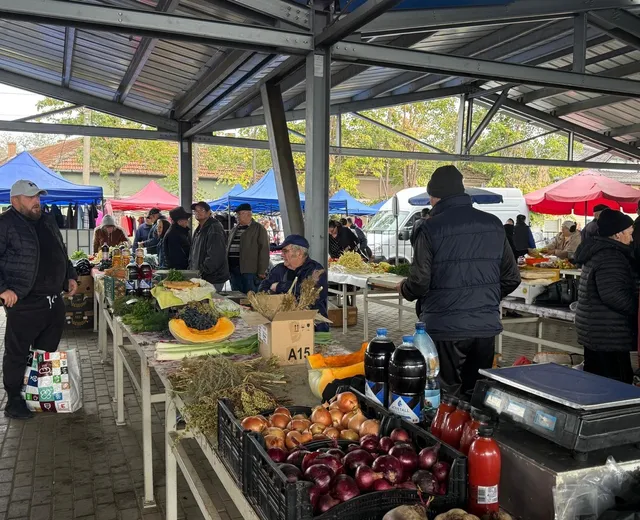O piață acoperită cu tablouri metalice, cu standuri pline cu legume, fructe și oameni care cumpără și vând.
