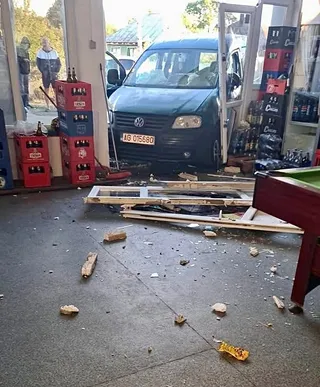 Un autoturism de culoare închisă este parcat într-un garaj lângă cutii de baterii și resturi de materiale pe podea.