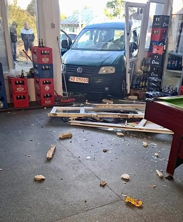 Un autoturism de culoare închisă este parcat într-un garaj lângă cutii de baterii și resturi de materiale pe podea.