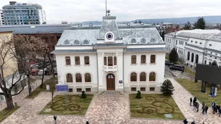 O vedere de sus a unei clădiri albe cu un acoperiș gri, situată lângă un drum cu mașini parcate și câteva persoane pe trotuar.