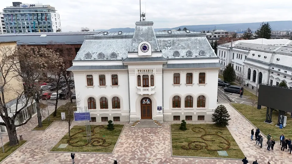 O vedere de sus a unei clădiri albe cu un acoperiș gri, situată lângă un drum cu mașini parcate și câteva persoane pe trotuar.