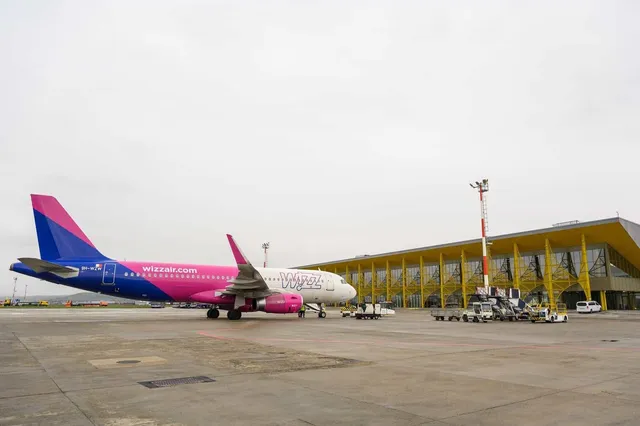 Un avion roz și alb cu inscripția wizzair.com stă pe o pistă de beton lângă un terminal de aeroport cu panouri galbene sub un cer înnorat.