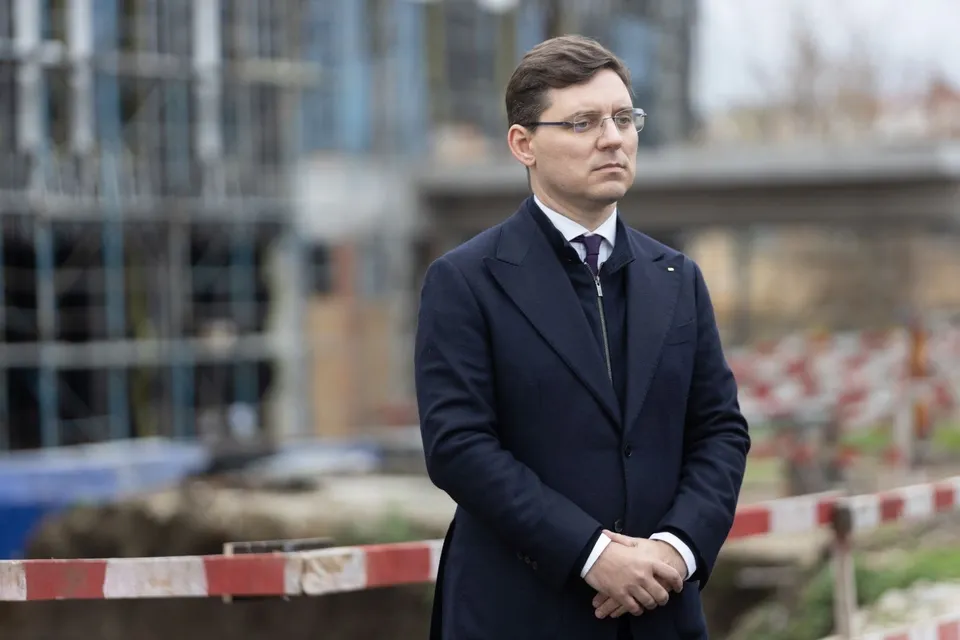 Un bărbat cu ochelari și costum închis la culoare stă cu mâinile încrucișate în față în fața unui fundal cu o clădire în construcție.