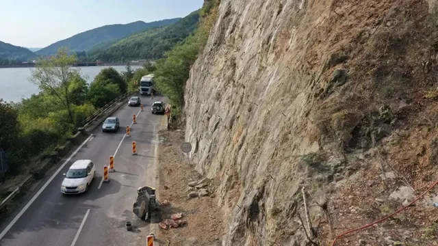 O stradă asfaltată cu mașini circulând pe lângă un versant stâncos și un corp de apă sub un cer senin.