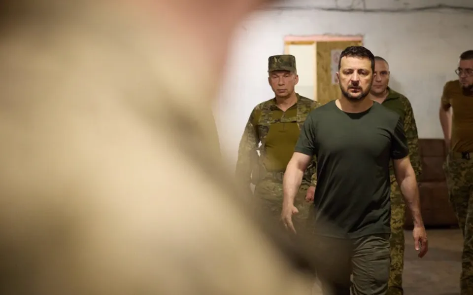 O imagine în plan apropiat cu un bărbat într-o tricou negru și pantaloni cargo, însoțit de mai multe persoane în uniformă militară, într-un interior cu pereți deschiși.