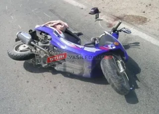 Un moped albastru este întins pe asfaltul unei străzi lângă o pată de lichid sau sânge.