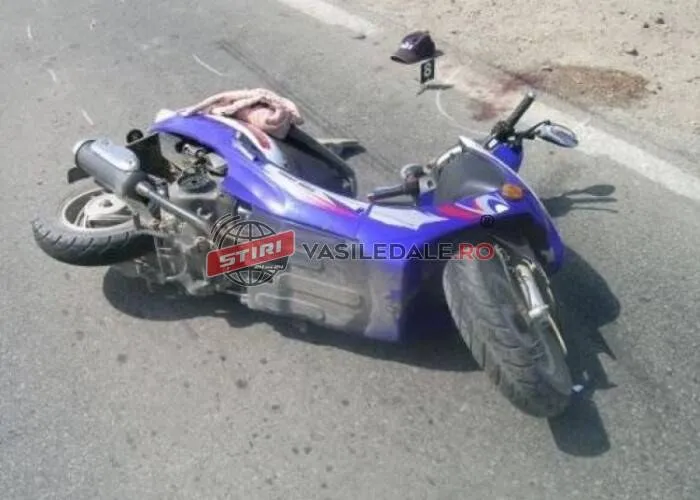 Un moped albastru este întins pe asfaltul unei străzi lângă o pată de lichid sau sânge.