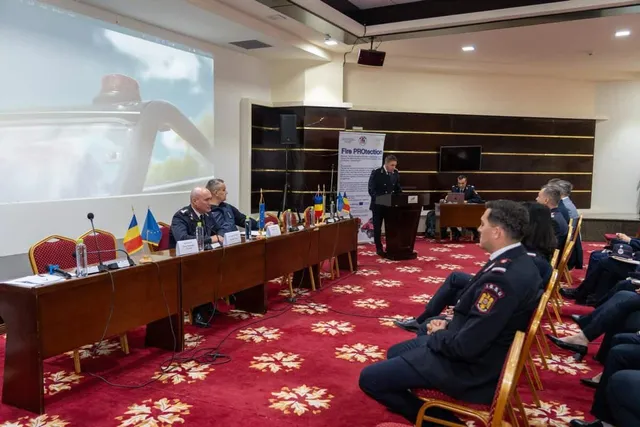 Oamenii în uniformă de serviciu stau la mese lungi într-o sală de conferințe cu covor roșu și modele florale, în timp ce un ecran proiectează o imagine.