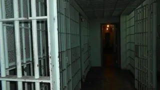 Un coridor întunecat este delimitat de celule de detenție cu gratii metalice de culoare deschisă.