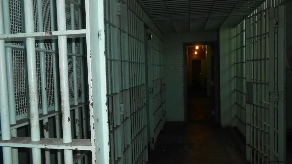 Un coridor întunecat este delimitat de celule de detenție cu gratii metalice de culoare deschisă.