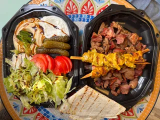 Două recipiente negre conțin mezze cu hummus, iaurt, castraveți murați, salată de roșii și verde, alături de fripturi pe băț și lipie.