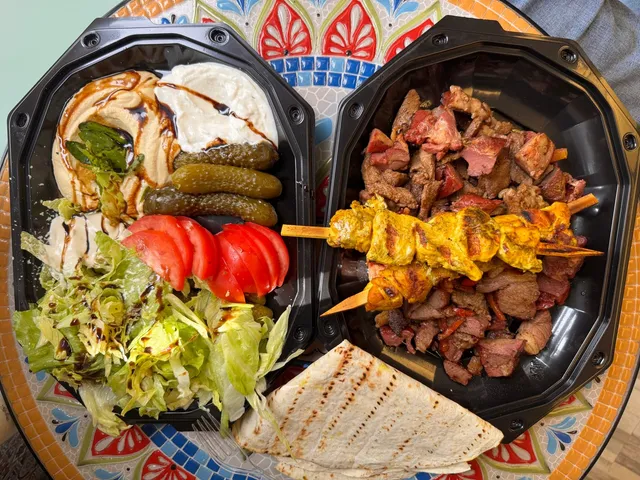 Două recipiente negre conțin mezze cu hummus, iaurt, castraveți murați, salată de roșii și verde, alături de fripturi pe băț și lipie.