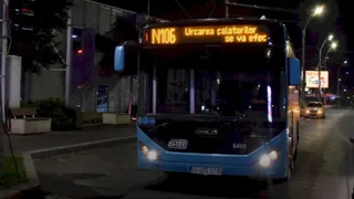 Un autobuz albastru cu afișaj digital portocaliu circulă pe un drum asfaltat noaptea, lângă un trotuar și un stâlp de iluminat stradal.