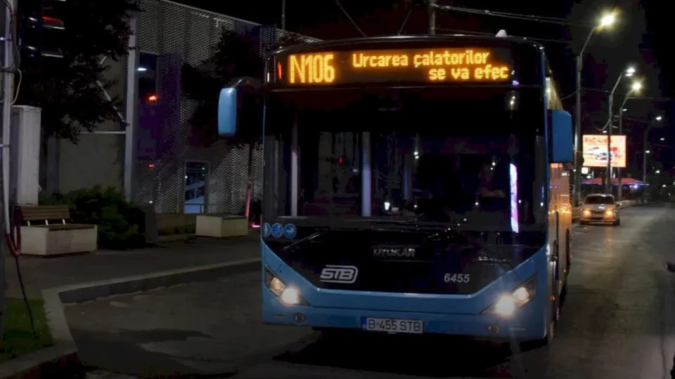 Un autobuz albastru cu afișaj digital portocaliu circulă pe un drum asfaltat noaptea, lângă un trotuar și un stâlp de iluminat stradal.