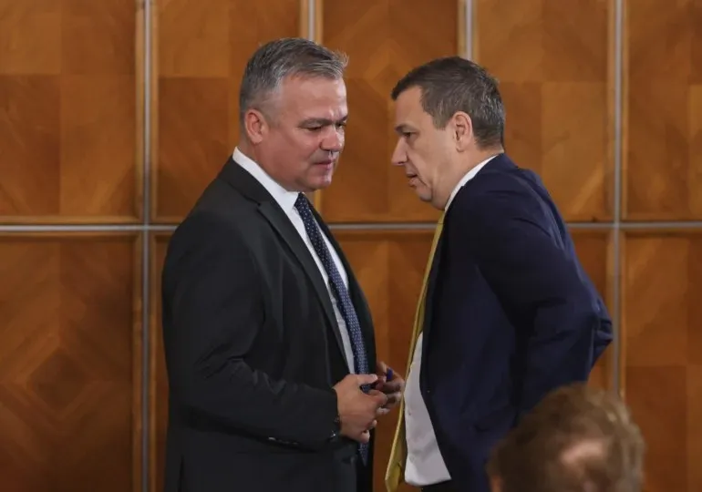 Doi bărbați în costume purtând cravate stau față în față în fața unui perete din panouri de lemn maro.