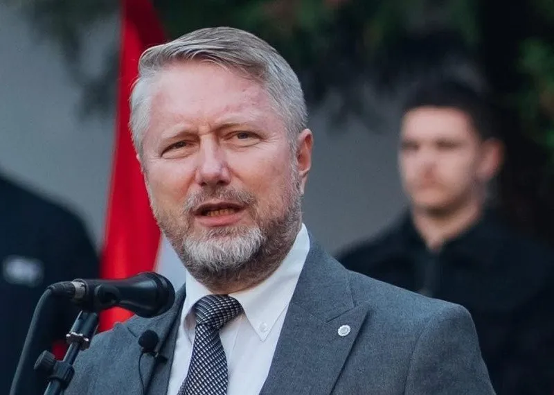 Un bărbat cu părul cărunt și barbă scurtă poartă un sacou gri, cămașă albă și cravată cu model, stând în fața unui microfon.