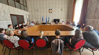Un grup de persoane stă așezat la o masă mare de lemn într-o încăpere cu pereți din lemn, orientate spre un ecran de televizor.