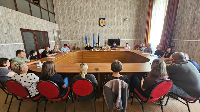 Un grup de persoane stă așezat la o masă mare de lemn într-o încăpere cu pereți din lemn, orientate spre un ecran de televizor.