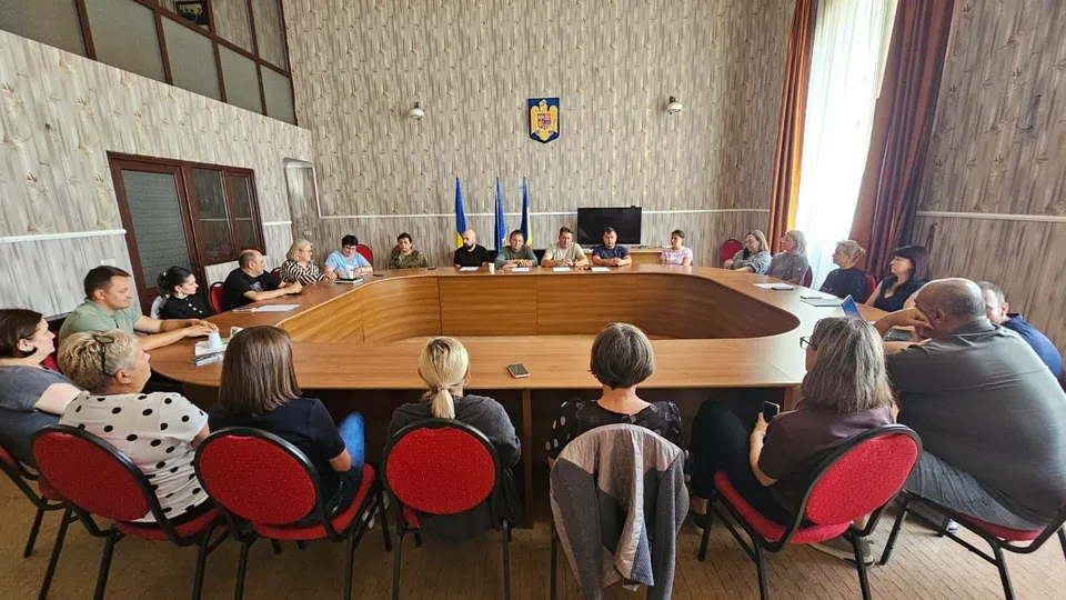 Un grup de persoane stă așezat la o masă mare de lemn într-o încăpere cu pereți din lemn, orientate spre un ecran de televizor.