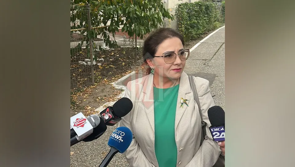 O femeie cu ochelari, purtând un sacou deschis peste un tricou verde, stă în fața mai multor microfoane întinse spre ea.