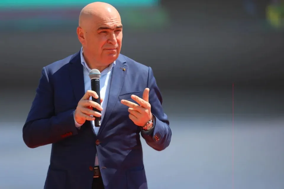 Un bărbat cu capul chel, purtând un sacou albastru și cămașă albă, ține un microfon și face un gest cu mâna dreaptă.