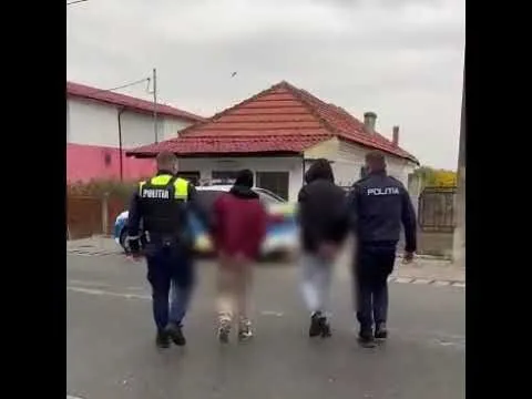 Patru persoane, dintre care doi polițiști în uniformă cu inscripția 'POLITIA', merg pe un drum spre un vehicul și o clădire cu acoperiș roșu.