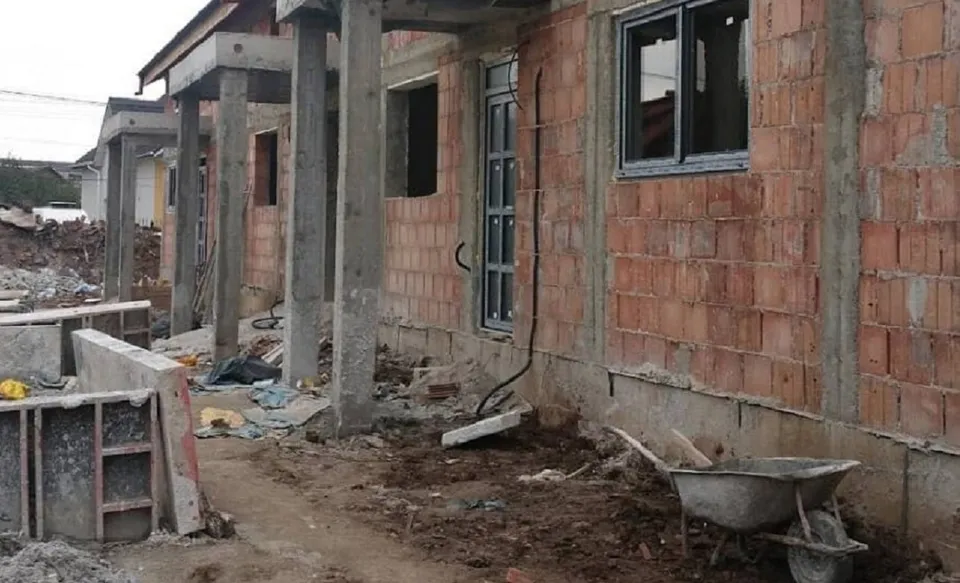 O clădire din cărămidă în construcție prezintă stâlpi de beton, ferestre negre, un cărucior de sârmă și resturi de materiale pe un teren de pământ.