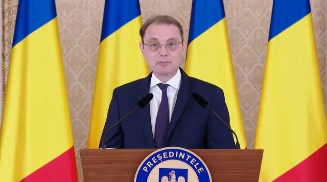 Un bărbat cu ochelari și cravată purtând un costum închis stă în spatele unui podium în fața a patru steaguri tricolore.