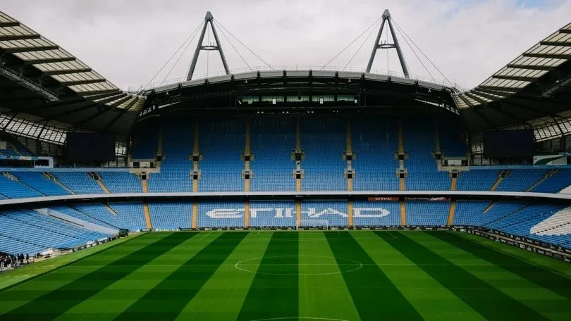 Un stadion de fotbal cu tribune de culoare albastră și un teren de gazon verde cu linii transversale sub un cer înnorat.