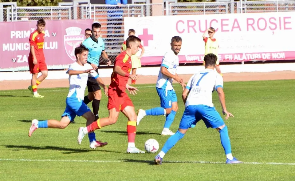 Câțiva jucători de fotbal în echipamente albastre și roșii dispută un meci pe un teren de iarbă verde sub lumină solară.